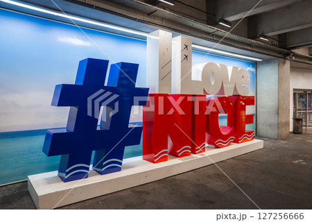 Indoor display of ILoveNice with blue, red, and white design, airplane icons, beach backdrop, and industrial airport setting in Nice, France. 127256666