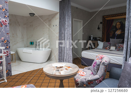 A cozy hotel room featuring a white bathtub in a marble alcove, floral wallpaper, plush furniture, a large bed, and a framed Mona Lisa portrait. 127256680