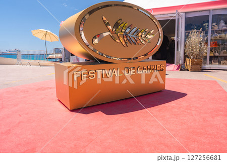 Golden Palme d'Or sculpture on red carpet with Festival de Cannes text, set outdoors with blue sky, parasol, waterfront, and nearby building. 127256681
