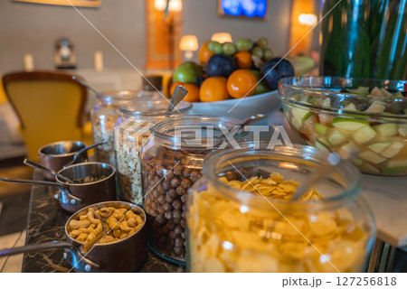 Marble countertop with glass jars of cereals, fresh fruit salad, and a decorative fruit platter. Yellow chair and soft lighting enhance the cozy ambiance. Marble countertop with glass jars of cereals, fresh fruit salad, and a decorative fruit platter. Yellow chair and soft lighting enhance the cozy ambiance. 127256818