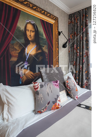 Hotel room featuring a framed Mona Lisa with sunglasses, white linens, floral pillows, floral curtains, and a modern wall mounted lamp. 127256820