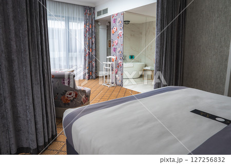 A cozy hotel room with a plush bed, floral armchair, open plan marble bathroom, freestanding bathtub, and soft lighting creating a warm ambiance. A cozy hotel room with a plush bed, floral armchair, open plan marble bathroom, freestanding bathtub, and soft lighting creating a warm ambiance. 127256832