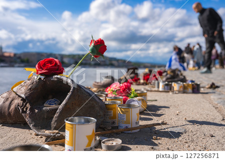 The Shoes on the Danube Bank memorial in Budapest features rusted iron shoes adorned with red roses and candles, with the Danube River in the background. 127256871