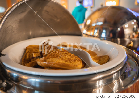 Close up of golden brown pancakes in a stainless steel chafing dish, with warm lighting, reflective surfaces, and a person in a teal hoodie in the background. Close up of golden brown pancakes in a stainless steel chafing dish, with warm lighting, reflective surfaces, and a person in a teal hoodie in the background. 127256906
