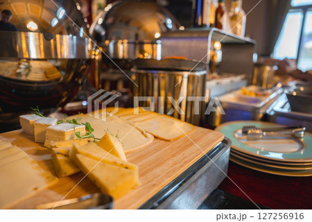An elegant cheese buffet on a wooden board with fresh herbs, metallic serving domes, and cutlery in a well lit boutique hotel dining area. An elegant cheese buffet on a wooden board with fresh herbs, metallic serving domes, and cutlery in a well lit boutique hotel dining area. 127256916