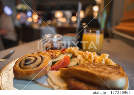 A cinnamon roll, toast, eggs, beans, and vegetables on a plate with orange juice. Warmly lit dining area with elegant decor and blurred figures in the background. 127256963