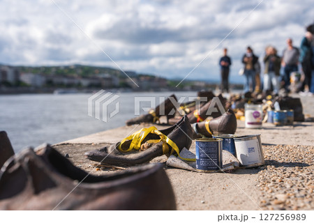 Iron shoes line the Danube River in Budapest, Hungary, with yellow ribbons and cans. Blurred figures and a cityscape appear under a partly cloudy sky. 127256989
