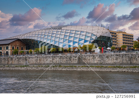 The Balna Budapest, a modern glass and steel building, stands by the Danube River with trees, a riverside promenade, and a partly cloudy sky. The Balna Budapest, a modern glass and steel building, stands by the Danube River with trees, a riverside promenade, and a partly cloudy sky. 127256991