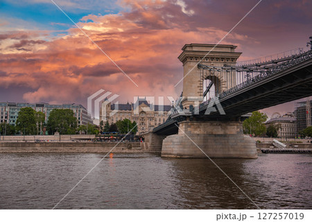 The Szechenyi Chain Bridge spans the Danube River in Budapest at sunset, with a vibrant sky, neo Gothic architecture, and people near the riverbank. The Szechenyi Chain Bridge spans the Danube River in Budapest at sunset, with a vibrant sky, neo Gothic architecture, and people near the riverbank. 127257019