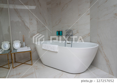 Elegant bathroom featuring a white freestanding bathtub, marble tiles with beige veining, gold framed side table, and decorative mirrors. Elegant bathroom featuring a white freestanding bathtub, marble tiles with beige veining, gold framed side table, and decorative mirrors. 127257052