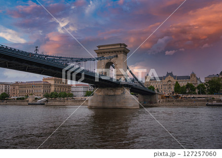 The Szechenyi Chain Bridge spans the Danube River in Budapest, Hungary, with stone towers, suspension cables, and a vibrant sunset sky reflected in the water. 127257060
