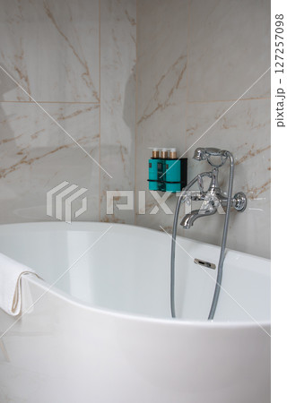 A modern bathroom featuring a white freestanding bathtub, chrome faucet, beige marble walls, and neatly arranged toiletries in a boutique hotel setting. A modern bathroom featuring a white freestanding bathtub, chrome faucet, beige marble walls, and neatly arranged toiletries in a boutique hotel setting. 127257098