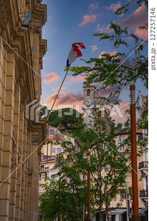 A street in Cannes, France, with a view of the Notre Dame de l'Esperance clock tower, French flag, classic architecture, and lush green trees. 127257146