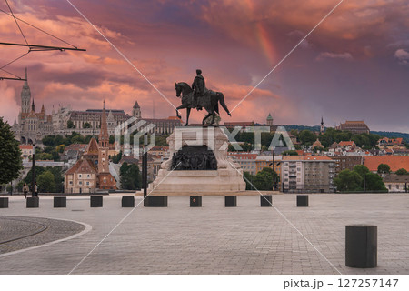 An equestrian statue in Budapest, Hungary, stands on a paved square with the Hungarian Parliament in the background under a sunset sky with a rainbow. 127257147