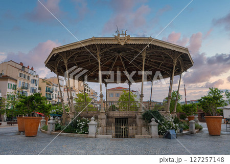 An octagonal bandstand with intricate ironwork and a decorative roof, surrounded by plants, trees, and pastel buildings in a cobblestone square in Cannes. An octagonal bandstand with intricate ironwork and a decorative roof, surrounded by plants, trees, and pastel buildings in a cobblestone square in Cannes. 127257148