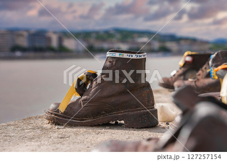 Bronze shoe sculpture with ribbon and Hebrew text at Shoes on the Danube Bank memorial in Budapest, Hungary, with Danube River in the background. Bronze shoe sculpture with ribbon and Hebrew text at Shoes on the Danube Bank memorial in Budapest, Hungary, with Danube River in the background. 127257154
