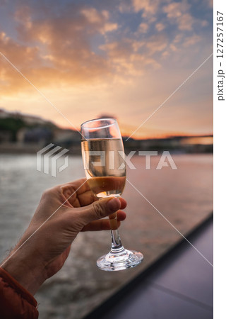 A hand holds a champagne glass by the Danube River at sunset in Budapest, Hungary. Soft orange and pink hues reflect on the water with buildings in view. 127257167