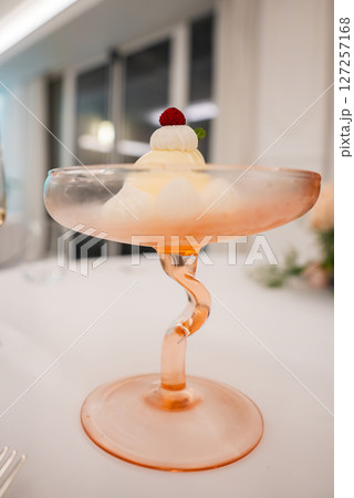 A creamy dessert served in a pink hued twisted stem glass, garnished with a red berry and mint leaf. Set in a modern dining space with large windows. A creamy dessert served in a pink hued twisted stem glass, garnished with a red berry and mint leaf. Set in a modern dining space with large windows. 127257168