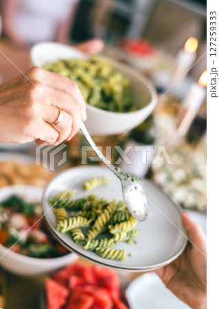 Close-up of hands serving green pesto pasta with spoon on white plate during festive summer meal 127259333
