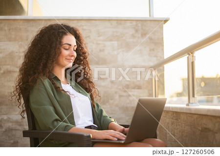 Beautiful curly woman working on laptop sitting on balcony at sunset, warm sun rays. Work on the Internet, online shopping, communication on social networks, online learning, freelancer, remote work Beautiful curly woman working on laptop sitting on balcony at sunset, warm sun rays. Work on the Internet, online shopping, communication on social networks, online learning, freelancer, remote work 127260506