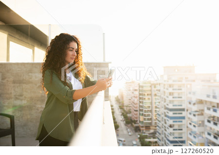 Young curly girl standing on the veranda overlooking the city with a phone in her hands, calm look, online work, online shopping, social networking, online learning 127260520