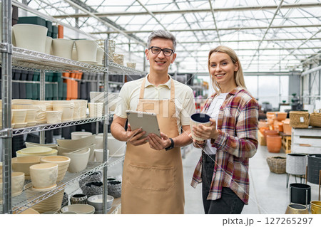 A shop employee helps a customer select flowerpots in a garden center, surrounded by various plant containers. A shop employee helps a customer select flowerpots in a garden center, surrounded by various plant containers. 127265297
