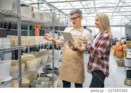 A Caucasian woman discusses flower pots with a shop assistant at a plant store. They're examining options on a tablet device near shelves. 127265302
