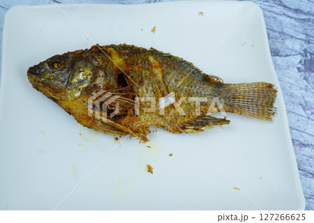 A perfectly golden-fried Mujair or Tilapia rests on a white rectangular plate, with a small bowl of dark dipping sauce in the background. A perfectly golden-fried Mujair or Tilapia rests on a white rectangular plate, with a small bowl of dark dipping sauce in the background. 127266625