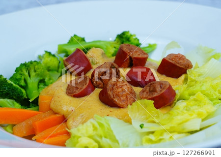A white plate holds cooked broccoli, cabbage, carrots, and sliced sausages atop a creamy yellow sauce, artfully arranged. 127266991