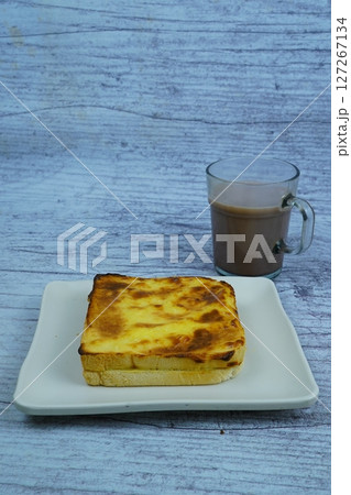A golden-brown baked toast with a creamy topping is served on a white plate next to a glass of chocolate milk, all on a rustic wooden background. A golden-brown baked toast with a creamy topping is served on a white plate next to a glass of chocolate milk, all on a rustic wooden background. 127267134