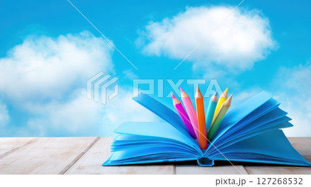 Open book with colorful pencils against a blue sky and clouds 127268532