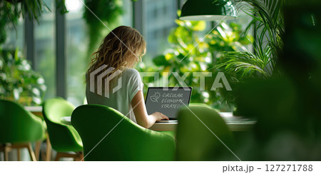 Woman working on laptop in green and white modern office filled with plants, focusing on circular economy strategies 127271788