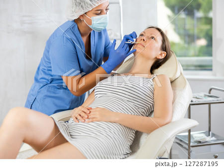 Doctor in disposable mask performs procedure of lip augmentation of girl patient in clinic. 127287195