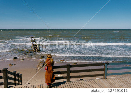woman in a brown coat stands on a pier overlooking the ocean and enjoys the views.  127291011