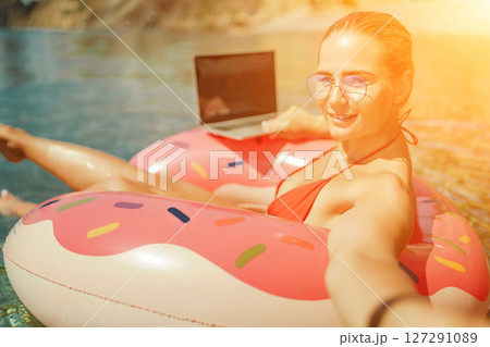 Woman laptop sea. Freelancer woman in sunglases floating on an inflatable big pink donut with a laptop in the sea. People summer vacation rest lifestyle concept 127291089