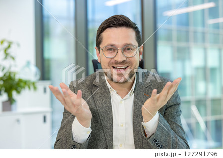 A young businessman in a suit and glasses sits in the office at a desk and talks to the camera while gesturing with his hands. 127291796