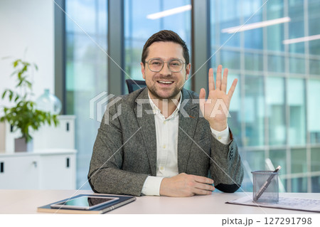 Smiling young man sitting at his workplace in the office, greeting and waving at the camera. Smiling young man sitting at his workplace in the office, greeting and waving at the camera. 127291798
