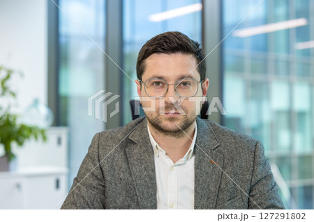 Portrait of a young male businessman sitting in the office at his workplace and looking seriously at the camera. Portrait of a young male businessman sitting in the office at his workplace and looking seriously at the camera. 127291802
