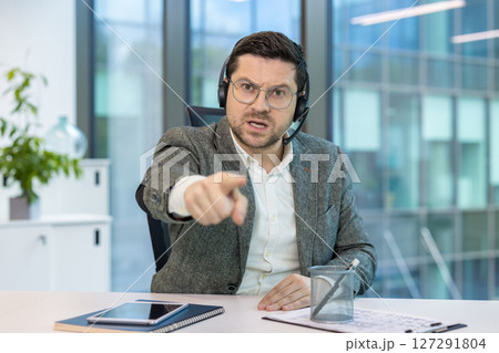 A man in a business suit, wearing a headset, points accusingly while looking directly at the camera. The office setting has natural light. A man in a business suit, wearing a headset, points accusingly while looking directly at the camera. The office setting has natural light. 127291804