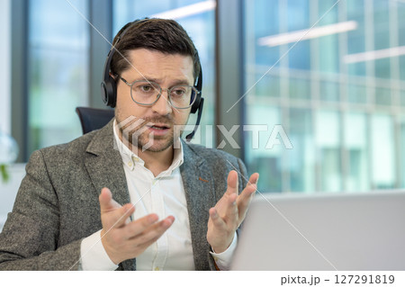 A man wearing a headset speaks in front of a laptop, appearing engaged in a virtual meeting at the office. A man wearing a headset speaks in front of a laptop, appearing engaged in a virtual meeting at the office. 127291819