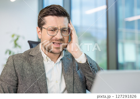 Upset young male businessman sitting in the office at his workplace, looking at the laptop screen, holding his head, solving a problem and making a mistake. 127291826