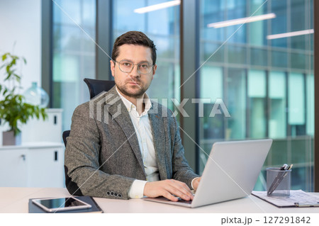 Portrait of a serious and confident young man in a suit and glasses sitting in the office at a desk, working on a laptop and looking at the camera. Portrait of a serious and confident young man in a suit and glasses sitting in the office at a desk, working on a laptop and looking at the camera. 127291842