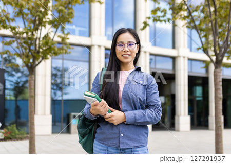 A smiling asian student, wearing glasses, standing in front of a university building with books in hand, carrying a backpack. A smiling asian student, wearing glasses, standing in front of a university building with books in hand, carrying a backpack. 127291937