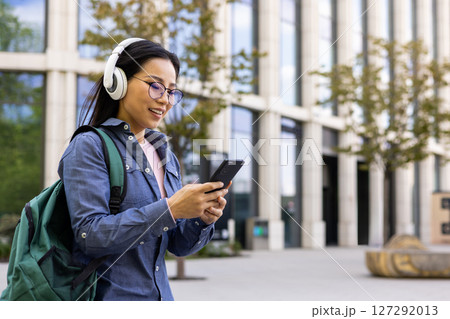 An asian woman walks and looks at her phone, wearing headphones and a backpack. She smiles while checking her smartphone in front of a modern building. 127292013