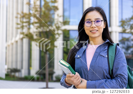 A smiling Asian woman stands in front of a modern building, holding books and a tablet with a backpack. A smiling Asian woman stands in front of a modern building, holding books and a tablet with a backpack. 127292033