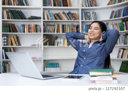A relaxed woman smiles with her eyes closed, enjoying a break in the library. Her arms rest behind her head as she sits at her desk. Bookshelves fill the background. A relaxed woman smiles with her eyes closed, enjoying a break in the library. Her arms rest behind her head as she sits at her desk. Bookshelves fill the background. 127292107