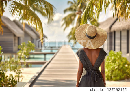 Woman in a stylish summer dress and wide-brimmed hat walks along a wooden pathway surrounded by tropical palm trees and beachside villas, embracing a serene summer escape 127292155
