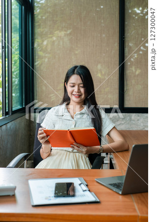 A young woman sits at a desk in a modern office, smiling while reading a tablet. Natural light filters through large windows. 127300047