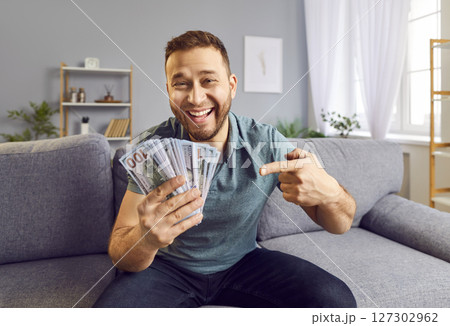 Happy excited young man pointing finger on fan of cash money dollar bills sitting on sofa at home 127302962
