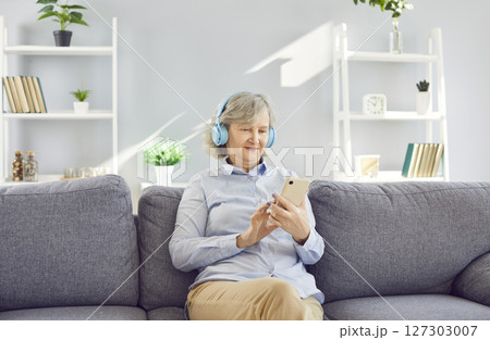 Senior woman listening to music with headphones using mobile phone at home. 127303007